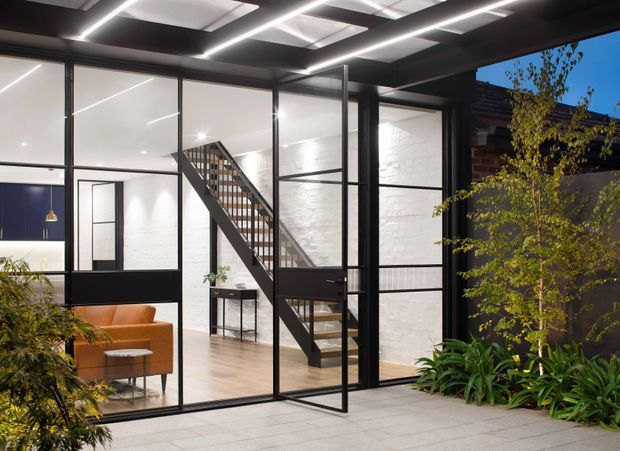 A contemporary glazed courtyard or entry view into a refined industrial-style interior. Black metal-framed glass partitions and doors reveal a timber staircase, pale timber flooring, white-painted brick walls, and a warm tan sofa. Linear recessed ceiling lighting and roof glazing create a bright, structured atmosphere, while planted landscaping frames the exterior foreground.