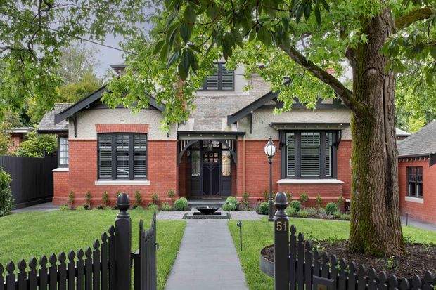 Front exterior view of a traditional brick residence, showing a symmetrical facade with contrasting rendered brickwork, dark gabled roof sections, black louvered shutters, a covered entry porch, a central walkway, manicured lawn, and a black picket fence beneath mature trees.