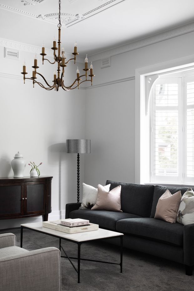 A refined living room with white walls, ornate ceiling cornices and decorative plaster detailing, a dark charcoal sofa with pale cushions, a dark timber sideboard, and a white rectangular coffee table. A traditional brass chandelier hangs prominently from the decorated ceiling, while plantation shutters frame the bright window. The space has a monochromatic, elegant and comfortably furnished appearance.