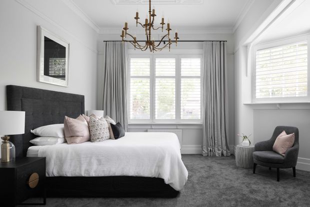 A refined bedroom with a large upholstered charcoal headboard and matching dark bed base, dressed in white bedding with blush and patterned accent cushions. The room has pale walls with decorative ceiling moulding, full-height grey curtains, plantation shutters, a classic brass chandelier, dark bedside furniture, and a grey carpet. A reading chair and small side table sit beside the window.