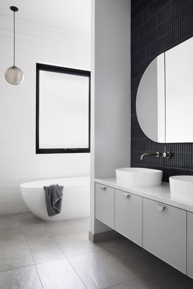 A bright contemporary bathroom featuring a freestanding white bathtub beside a black-framed window, a floating vanity with two white vessel basins, and a dramatic black vertically ribbed tiled feature wall with a large round mirror. The room uses a restrained monochrome palette with grey tiled flooring, white wall tiles, black metal details, and a spherical pendant light.