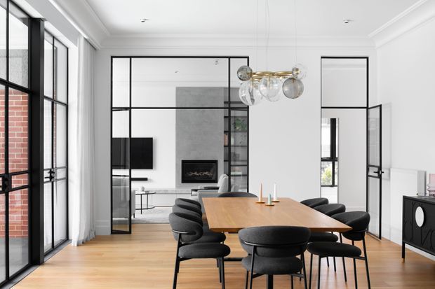 Bright, refined open-plan dining and living space with a large timber dining table surrounded by black upholstered chairs. Black-framed glazed partitions and doors define the room, with a contemporary fireplace, television, sculptural glass-and-metal pendant light, and a black sideboard. White walls, decorative ceiling moulding, and pale timber flooring create a high-contrast modern interior.