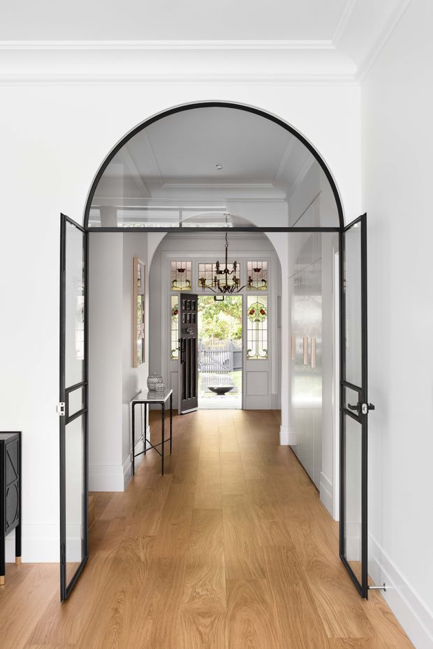 A bright, refined residential entry hall viewed through a dramatic black-framed arched glass doorway. The hallway features warm timber flooring, crisp white walls and ceilings with layered decorative moulding, white built-in cabinetry, a slim console table, and a traditional front door with stained-glass sidelights and an upper transom. A chandelier and garden view are visible beyond the entry.
