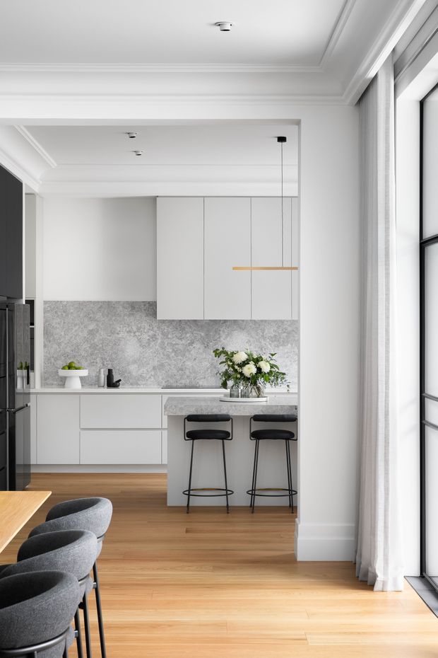 Bright, refined open-plan kitchen and dining area with white shaker-style cabinetry, a mottled grey stone-look splashback, pale timber flooring, and a compact island with two black stools. The space features ornate white ceiling cornices and recessed lighting, large black-framed windows with sheer curtains, and a partial dining table with upholstered grey chairs in the foreground. A linear brass pendant hangs above the island.