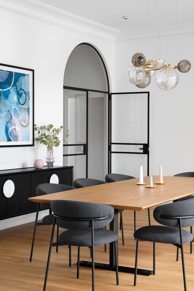 A refined dining room with a large rectangular natural timber dining table surrounded by charcoal upholstered chairs. The room has warm timber flooring, white walls with decorative ceiling cornices, black-framed glazed internal doors, a tall arched opening, a black sideboard with circular white inlays, abstract blue artwork, and a sculptural brass-and-glass globe pendant light.