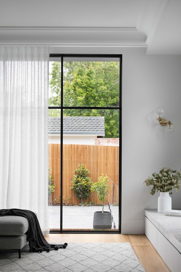 A bright, refined living room interior featuring a tall black-framed glazed door or window overlooking a private paved courtyard. The room has white walls and ornate stepped ceiling detailing, sheer white curtains, pale timber flooring, a grey upholstered sofa with a dark throw, a light patterned rug, and streamlined white built-in cabinetry with a pale stone-like top. A small brass-and-glass wall sconce and simple vase styling add subtle accents.