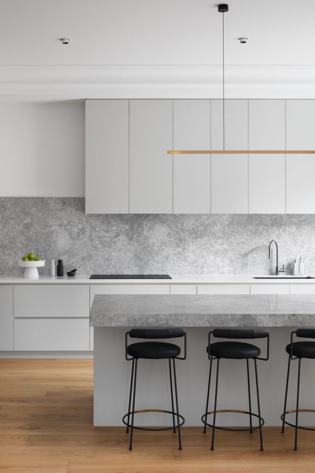 A refined contemporary kitchen with full-height matte white cabinetry, a grey textured stone splashback, and a large grey stone island with integrated seating. Three black bar stools sit at the island, while a slim linear pendant light provides a warm brass-toned accent. Light timber flooring adds warmth to the otherwise monochromatic space.
