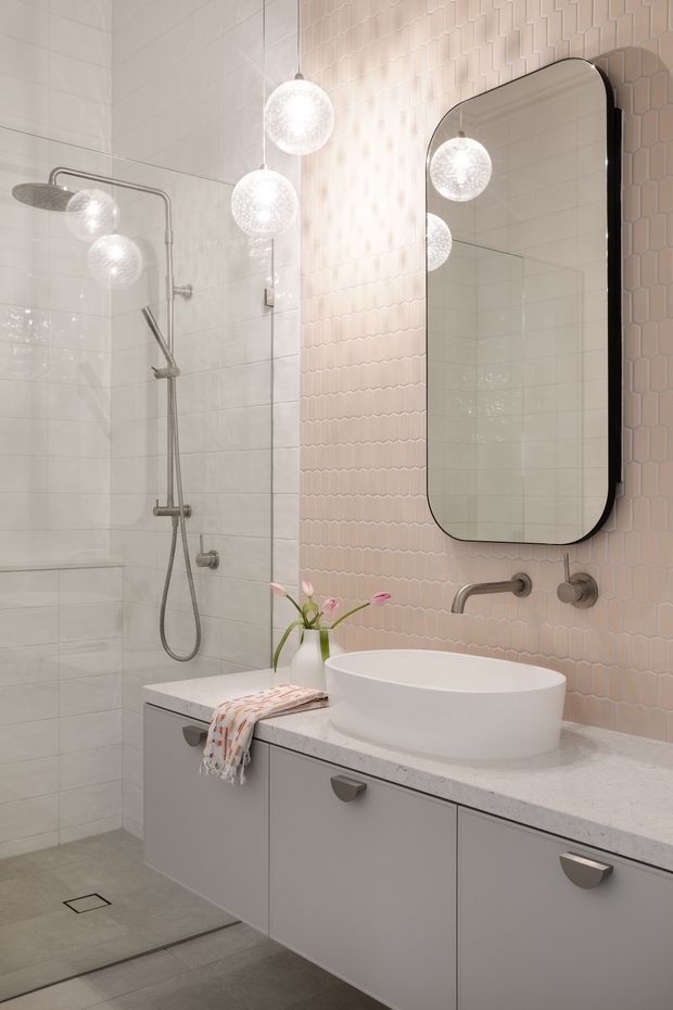 A contemporary bathroom with a walk-in shower, wall-mounted brushed metal fixtures, a floating grey vanity with a pale stone benchtop, and a white vessel basin. The walls combine large pale grey tiles with a blush-pink geometric feature tile, complemented by a rounded-rectangle black-framed mirror and spherical pendant lights. The room has a refined, softly illuminated aesthetic.