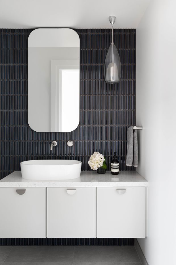 A contemporary bathroom vanity with a white vessel basin set on a pale stone benchtop, white flat-panel cabinetry, and a dramatic dark vertically ribbed tile feature wall. The wall includes a rounded-rectangle mirror, wall-mounted tapware, pendant light, and towel rail. Large grey floor tiles and restrained monochrome styling create a refined, modern look.