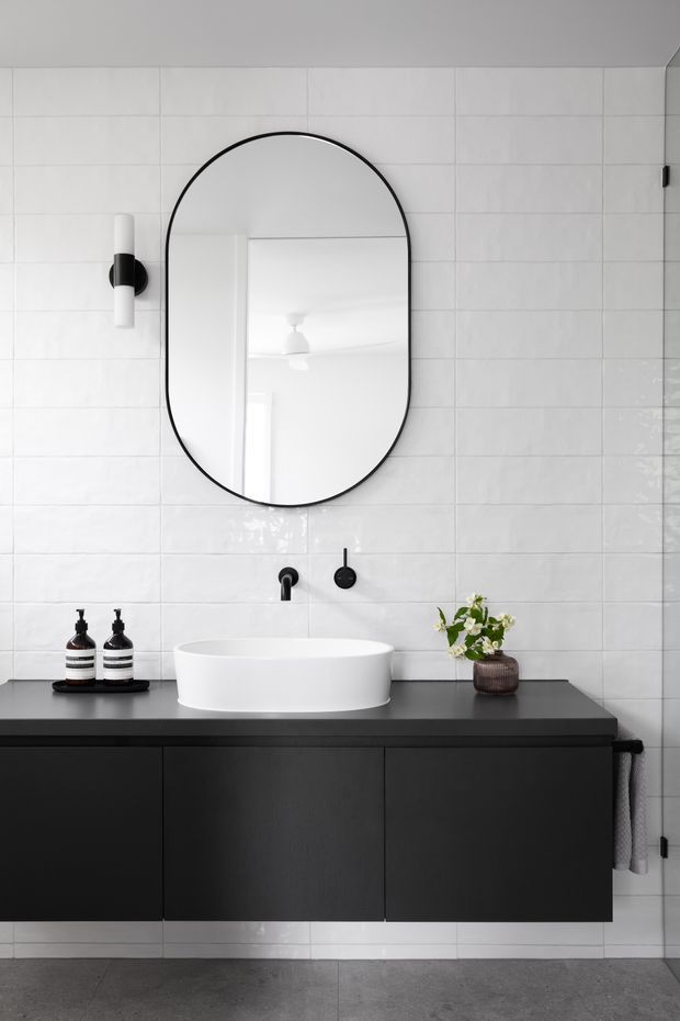 A contemporary bathroom vanity with a large black-framed rounded-rectangle mirror, white wall tiles, matte black floating cabinetry and benchtop, a white vessel basin, black wall-mounted tapware, and minimalist black-and-white accessories. The space has a clean, monochromatic, refined appearance with grey floor tiles.
