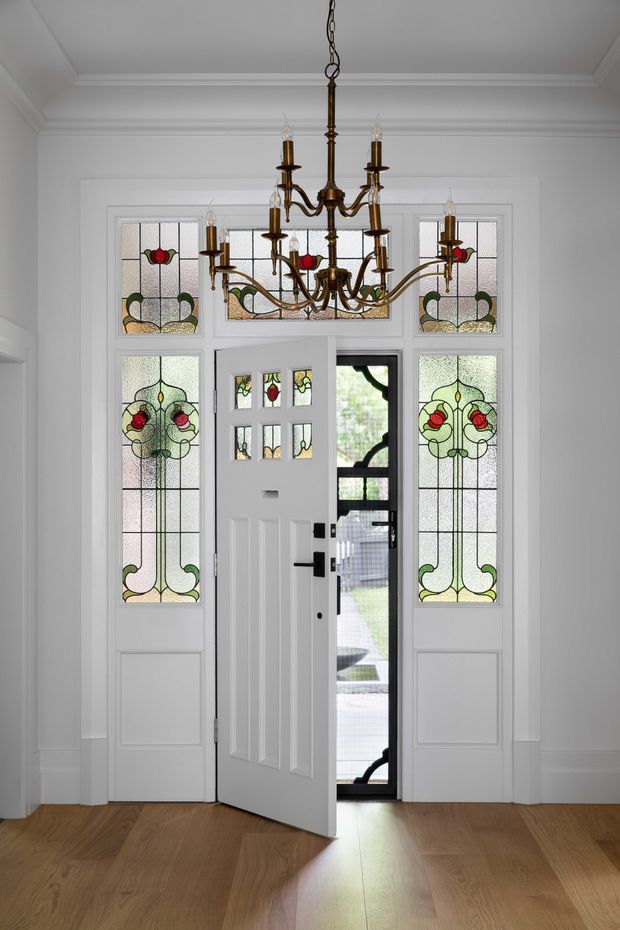 A bright, formal residential entryway viewed from inside. A white paneled front door is open within a symmetrical arrangement of decorative stained-glass sidelights and transom panels featuring floral Art Nouveau-style motifs. A substantial antique brass chandelier hangs from a high white coffered and moulded ceiling. White walls, ornate trim, black door hardware, and warm timber flooring create a refined heritage character.