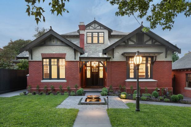 Front exterior view of a renovated or carefully maintained early-20th-century brick house, featuring a symmetrical gabled facade, dark timber Tudor-style detailing, rendered upper walls, slate-like roofing, multi-pane windows, and a covered central entry. A formal front garden with clipped planting, paving, and a small square water feature leads to the front door, while a traditional lamp post stands in the foreground.