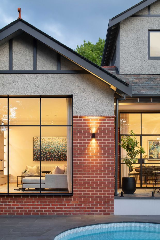 Contemporary residential exterior featuring steep gabled roof forms, dark metal framing and trim, textured light-grey render, and red brickwork. Large black-framed glazed openings reveal a warm living room and dining area, while a paved terrace borders the curved edge of a swimming pool in the foreground.