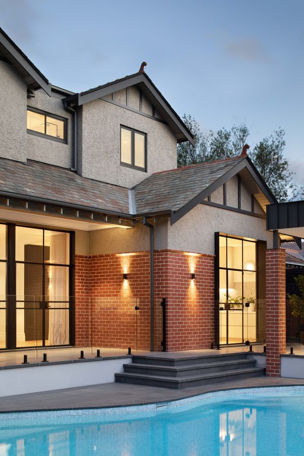 Exterior architectural photograph of a multi-level suburban house at dusk, focused on a brick-and-textured-render courtyard facade with steep gabled roof forms, dark-framed glazing, and a stepped terrace beside a swimming pool. Warm interior lighting and wall sconces contrast with the cool blue evening sky.