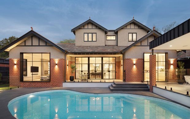 A large multi-wing suburban residence viewed from the pool, featuring red-brick walls, pale rendered upper sections, multiple steep gabled roofs with dark trim and decorative timber detailing, extensive glazed living areas, and a covered terrace beside a swimming pool. The image is a polished architectural exterior photograph at dusk.