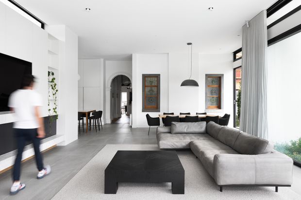 Bright, open-plan contemporary living space with a large grey sectional sofa, black geometric coffee table, wall-mounted television and dining area. White walls, grey tiled flooring, tall glazing with sheer curtains, arched doorway and decorative stained-glass-style windows create a clean, refined interior.
