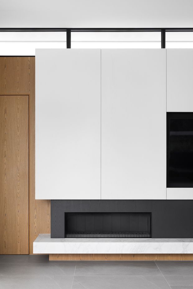 A close-up of a minimalist built-in living room media and fireplace wall. Warm timber veneer cabinetry and a concealed door sit beside large matte white overhead cabinets, with a black recessed fireplace and television niche below. A white marble-look hearth projects over dark charcoal wall panelling, and large grey floor tiles complete the restrained contemporary composition.