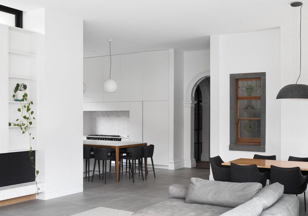 A bright open-plan interior combining a minimalist white kitchen, dining area, and living room. The kitchen has full-height white cabinetry, a dark benchtop, and a timber dining table with black chairs. A grey sectional sofa occupies the foreground, while a decorative arched doorway and timber-framed stained-glass window add traditional architectural detailing to the otherwise contemporary space.