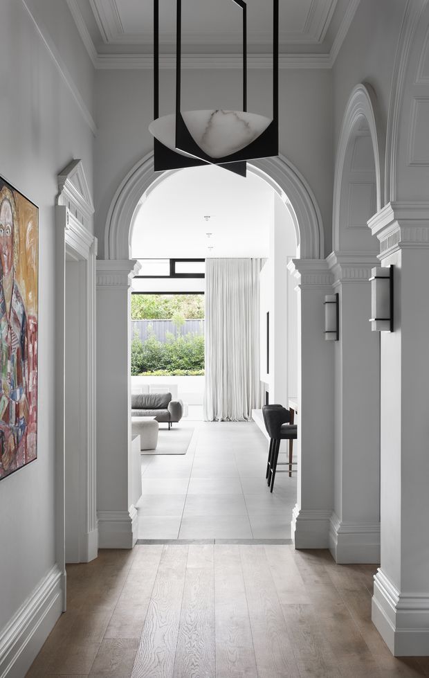 A refined interior hallway viewed toward an open-plan living area, framed by ornate white-painted architraves, pilasters, cornices and arched openings. The hallway has a pale timber floor, grey-white walls, contemporary black wall sconces, and a striking geometric pendant with a white marble-like shade. Beyond the arch is a bright living and dining space with full-height curtains, pale tiled flooring, modern seating and garden views.