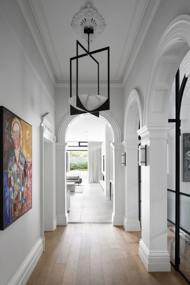 A refined, light-filled residential hallway with pale timber flooring, white paneled walls, ornate ceiling cornices, decorative archways, classical columns and elaborate mouldings. A striking black geometric pendant light hangs centrally, while colourful artwork lines the left wall and black-framed glazed doors or partitions appear to the right. The hallway opens toward a bright living area beyond.