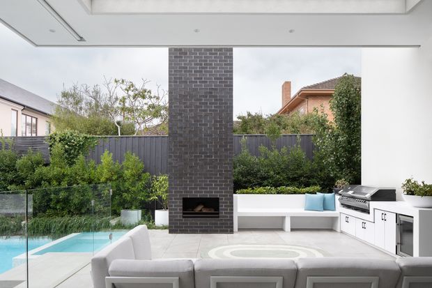 A bright contemporary outdoor entertaining area viewed from inside or beneath a covered pavilion, with a white built-in outdoor kitchen and cabinetry, long built-in bench seating, a dark brick fireplace, pool glimpses, and a large light-coloured sectional sofa. The space opens onto a landscaped suburban garden through expansive glazing.