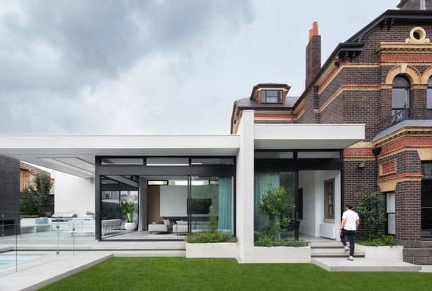 Exterior view of a contemporary white and glass extension attached to a historic dark-brick, ornate masonry residence. The extension features a flat roof, broad black-framed glazing, landscaped planter beds, a paved terrace and a glazed pool edge, creating a strong contrast between modern addition and heritage facade.