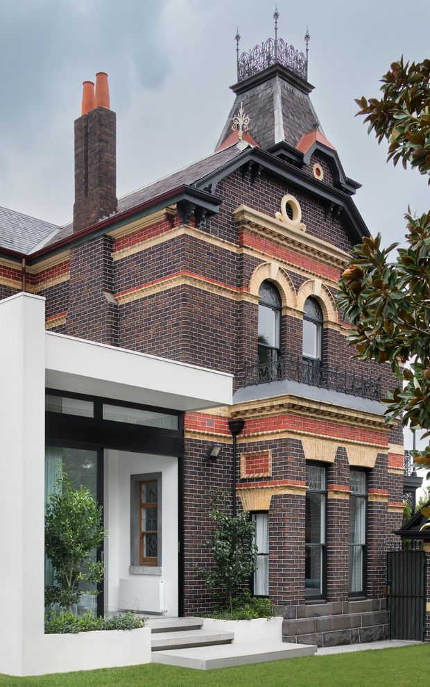 Exterior architectural photograph of a substantial historic dark brick building with ornate polychromatic brick detailing, arched windows, decorative stone bands, an elaborate slate-clad tower roof with finials, and a tall brick chimney. A crisp white contemporary glazed entrance extension projects from the lower left, creating a clear heritage-and-modern addition contrast. Landscaping and a lawn frame the foreground.