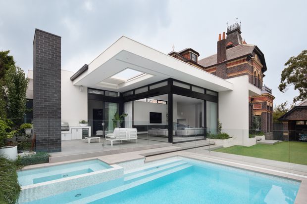 Contemporary flat-roof extension with crisp white walls, extensive black-framed glazing and a covered outdoor living area, set beside an ornate heritage brick residence. The image shows a bright turquoise swimming pool, raised spa, paved terrace, lawn and landscaped edges.