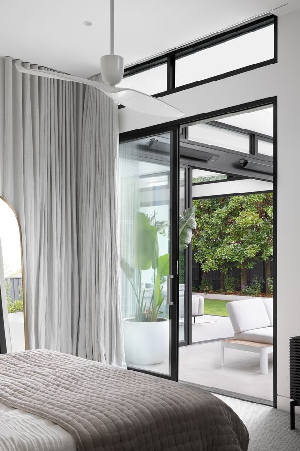 Bright contemporary bedroom opening directly onto a landscaped courtyard through large black-framed glazed doors. The room features a quilted neutral bedspread, full-height pleated curtains, a sculptural white ceiling fan, a tall indoor planter, and a white lounge chair visible outside. High-level clerestory glazing and strong black window framing contribute to an airy, minimalist composition.