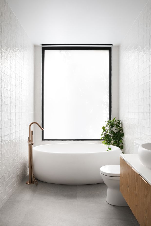 A bright, narrow contemporary bathroom featuring a freestanding white bathtub beneath a large black-framed frosted window, white square wall tiles, large light-grey floor tiles, a wall-mounted timber vanity, white toilet, and a brushed bronze floor-mounted bath tap. A leafy plant adds a natural accent.