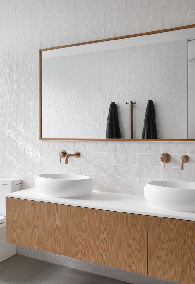 A bright contemporary bathroom featuring a wall-to-wall rectangular mirror with a timber frame, two white vessel basins on a floating timber vanity, wall-mounted brushed bronze taps, and white textured rectangular wall tiles. A toilet is partially visible at left, with a grey tiled floor and dark towels reflected in the mirror.