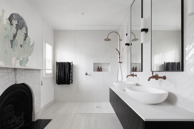 Bright, refined contemporary bathroom featuring a double vanity with vessel basins, wall-mounted bronze-toned tapware, a frameless glass shower enclosure, white herringbone wall tiles, pale timber-look flooring, dark vanity cabinetry, and a marble-clad fireplace surround with artwork.