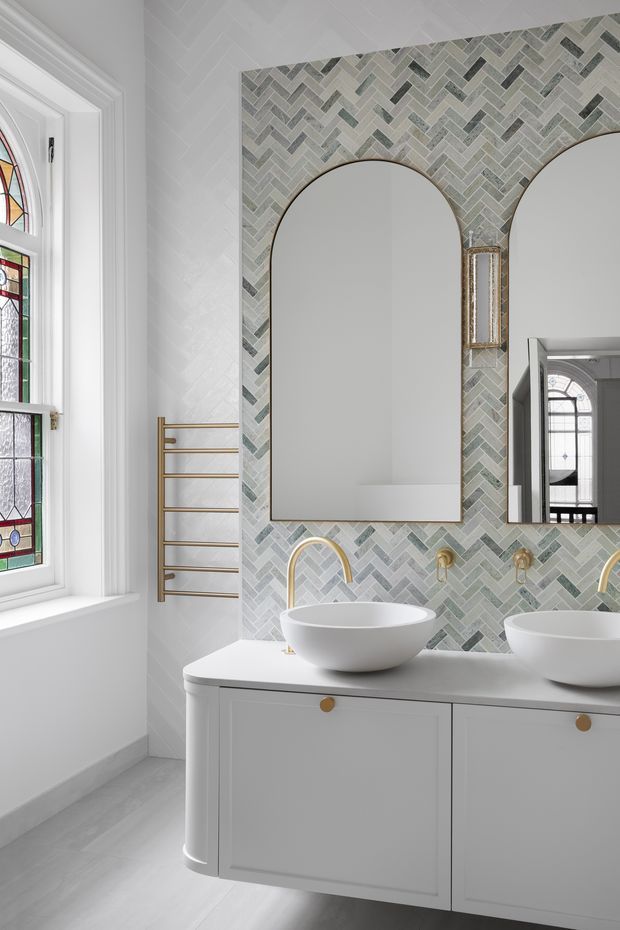 Bright, refined bathroom interior featuring a white double vanity with vessel basins, brushed brass wall-mounted faucets, arched mirrors, and a large geometric herringbone-pattern tiled feature wall in pale grey, white, and muted green tones. A stained-glass window and heated towel rail are visible on the left, with an additional mirrored view into the adjoining space on the right.