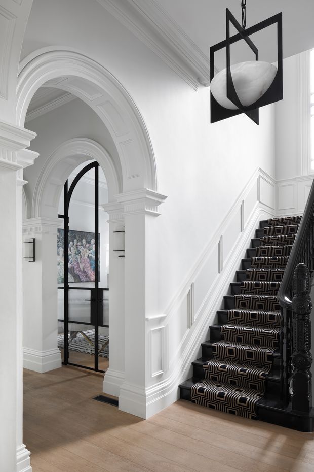 A refined residential entry hall featuring a grand white staircase with ornate black carved balustrade and patterned black-and-white stair runner. White architectural panelling, elaborate cornices, and repeated arched openings create a classic formal backdrop. A black-framed glazed door leads to a room with artwork beyond, while a sculptural black geometric pendant with a white stone-like centre hangs overhead. Light timber flooring balances the monochromatic scheme.