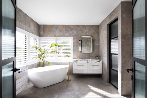 Bright contemporary bathroom with full-height grey stone-look wall and floor finishes, a freestanding white bathtub, white double vanity with integrated basins, brushed brass tapware, rounded rectangular mirror, and black-framed ribbed-glass doors. White plantation shutters and a potted palm add natural light and contrast.