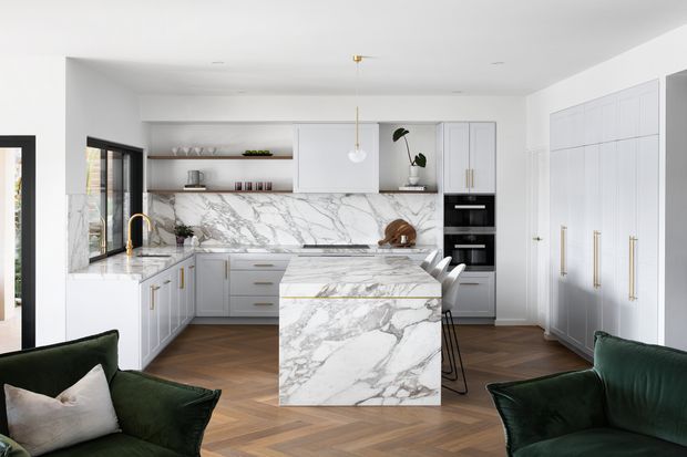 Bright, refined contemporary kitchen with white built-in cabinetry, a large marble-look waterfall island and matching backsplash, integrated black appliances, brass hardware and tapware, and warm herringbone timber flooring. Dark green lounge chairs in the foreground add contrast to the otherwise light, neutral scheme.