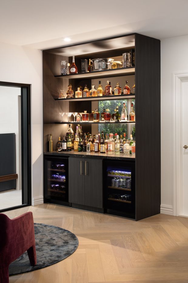 A refined built-in home bar alcove with dark timber cabinetry, open illuminated shelves displaying bottles and glassware, and integrated under-counter beverage or wine refrigerators. The bar is set against white walls with decorative trim and a light herringbone timber floor, adjacent to a living area.