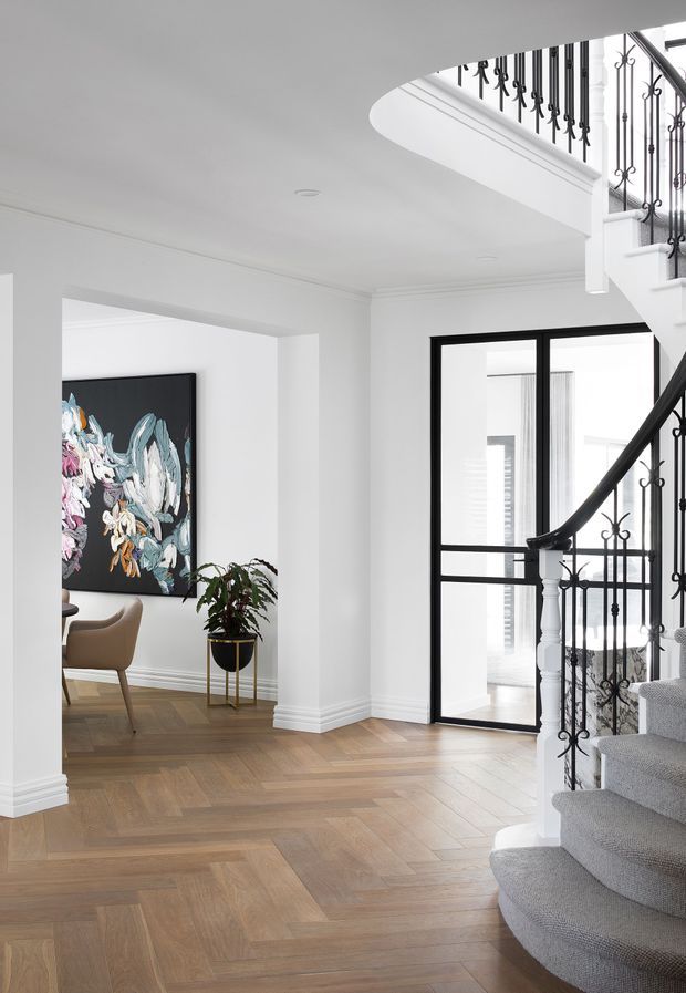 Bright, refined residential entry hall with a sweeping curved staircase, grey carpeted treads, ornate black metal balustrades, white walls and trim, and warm herringbone timber flooring. A black-framed glazed door opens toward a bright adjoining space, while a nearby sitting or dining area contains a contemporary abstract artwork, curved chair, and potted plant.
