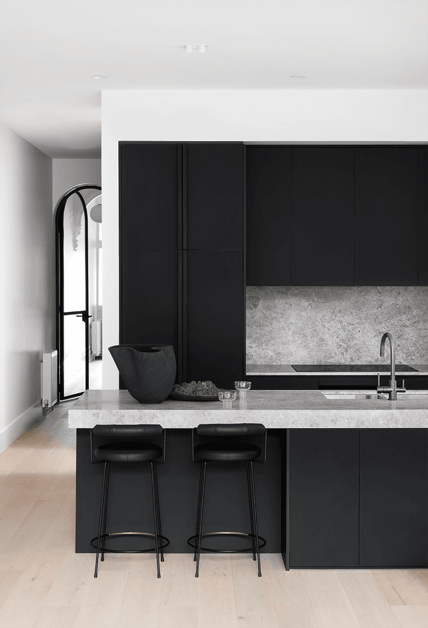A refined contemporary kitchen with a long island featuring a pale stone benchtop, integrated sink and tall curved tap. Matte-black full-height cabinetry and island joinery contrast with a textured grey stone splashback. Two black upholstered bar stools sit at the island, while a tall arched black-framed doorway or mirror is visible to the left. The room has pale timber flooring, white walls and ceiling, and a restrained monochromatic palette.