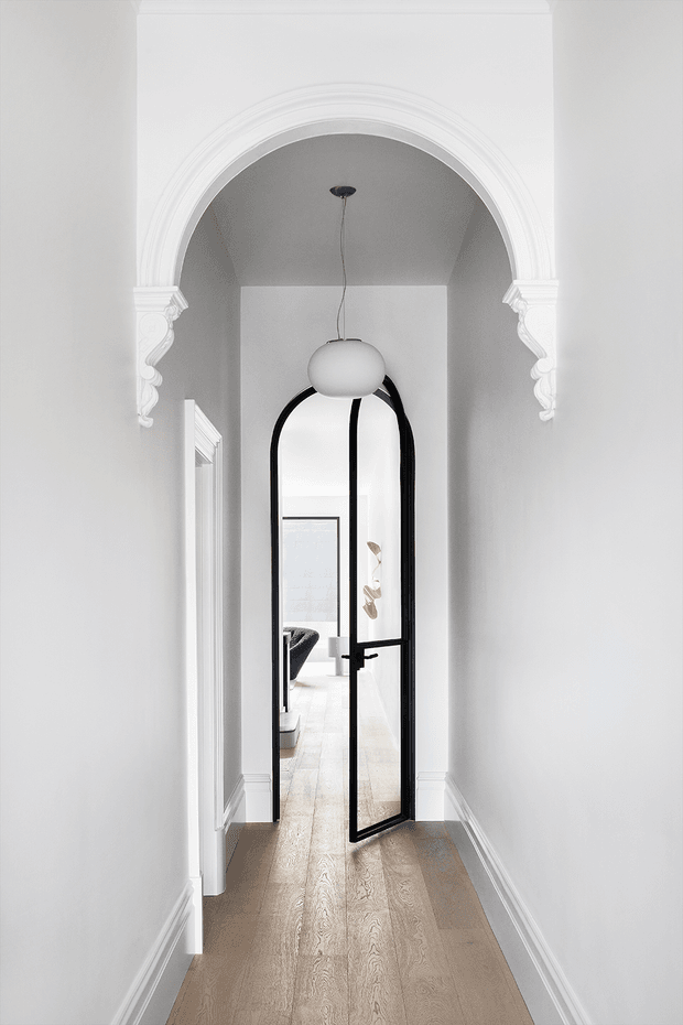 Bright, narrow residential hallway with pale timber flooring and white walls, framed by an ornate white decorative archway with carved corbels. A black-framed arched glass door stands open toward a light-filled room beyond, with a sculptural pendant light suspended in the passage. Traditional architectural detailing is contrasted with contemporary black metal glazing.