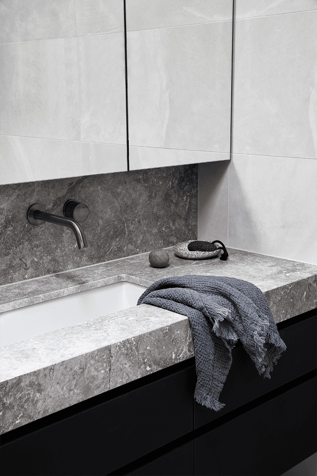 A close-up of a refined modern bathroom vanity featuring a deep integrated rectangular basin, dark stone-look countertop and backsplash, large pale stone wall tiles, matte black cabinetry, and a wall-mounted brushed-dark-metal tap. A textured blue-grey towel and small decorative accessories add contrast.