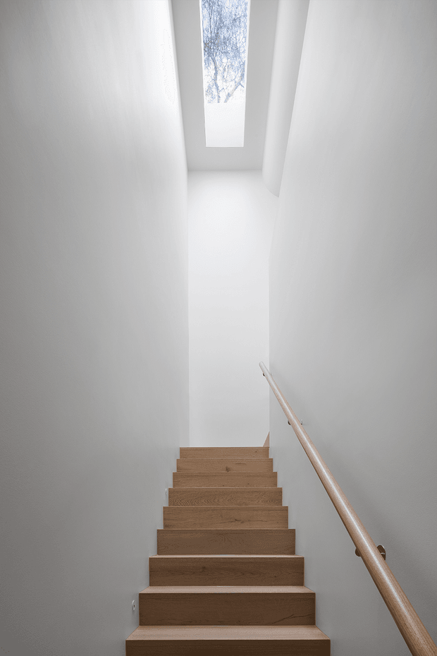 A narrow, minimalist stairwell viewed from the bottom, with a straight flight of warm natural timber stairs, smooth white walls, a simple timber handrail, and a tall skylight bringing in daylight from above. The space is clean, bright, and vertically elongated.