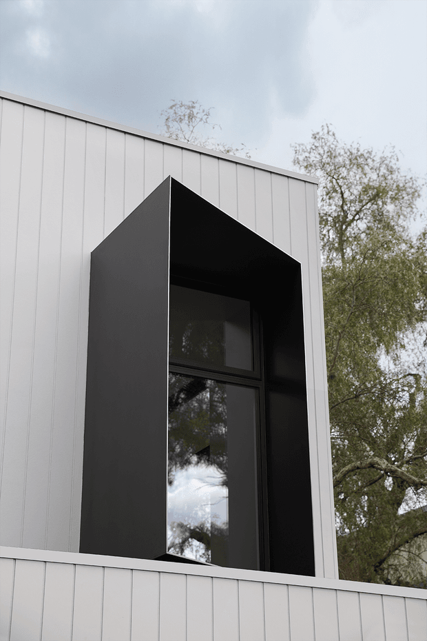 Close-up exterior view of a contemporary building facade clad in light vertical metal panels. A tall, deeply recessed black-framed window is surrounded by a projecting angular black metal reveal, creating a strong geometric feature. Mature trees are visible around the building, with an overcast sky in the background.