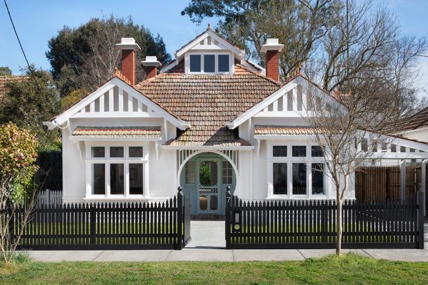 Front exterior view of a symmetrical heritage-style single-storey house with a prominent tiled pitched roof, multiple brick chimneys, white rendered walls, decorative gable timber detailing, and a central arched entry porch. A black picket fence and landscaped frontage frame the facade.