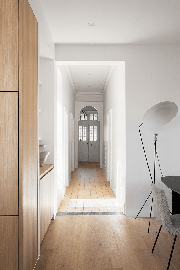 A bright, refined residential hallway viewed from an open-plan room. The space features pale timber flooring, white walls and detailed ceiling cornices, tall white skirting boards, a long corridor leading to a glazed entry door, and built-in light timber cabinetry along the left wall. A sculptural floor lamp and partial dining or work table with chair sit in the foreground.