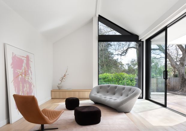 Bright contemporary living room with a sculptural light-grey tufted sofa, curved brown lounge chair, dark ottomans, pale timber flooring, and minimalist timber storage. Large black-framed glazed doors open onto an outdoor deck and leafy garden, while a large pink abstract artwork leans against the wall.
