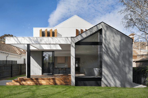 A contemporary residential extension viewed from the backyard, combining a dark grey gabled volume with extensive glazing and a white flat-roofed upper structure. The home features vertical cladding, timber screening elements, a covered outdoor deck, and strong geometric forms, set among neighbouring suburban houses and lawn.