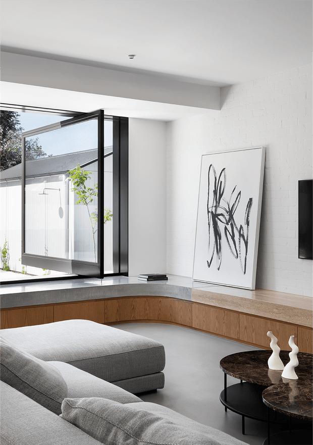 A bright contemporary living room with a large grey sectional sofa, curved built-in timber bench cabinetry, a dark stone-topped coffee table, and a black-framed folding glass door opening to an exterior courtyard. White-painted brick walls, exposed concrete-toned surfaces, minimalist abstract artwork, and sculptural white ceramics create a clean, refined interior.