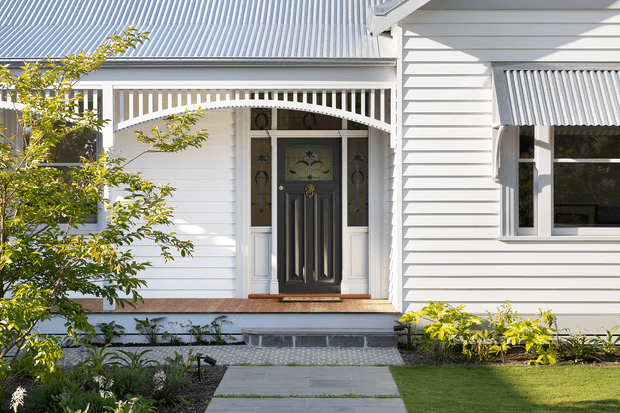 Front exterior view of a renovated white weatherboard house, featuring a corrugated metal roof, decorative timber porch balustrade and arch, ornate dark entry door with sidelights, and a landscaped garden with a concrete path.
