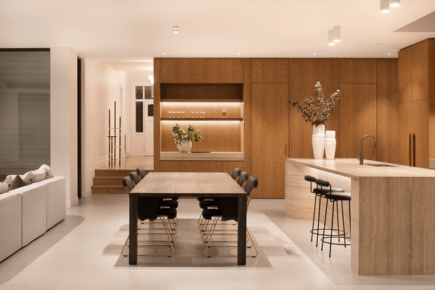 A warm, open-plan contemporary interior combining a dining area and kitchen. A dark wood dining table with black chairs sits on a pale rug, while a large light stone island with integrated seating and a sink occupies the right side. Full-height timber cabinetry and wall panelling form a feature backdrop, with illuminated open shelving displaying glassware and decor. Pale flooring, recessed lighting, and a glimpse of a living area and entry stairway complete the refined space.